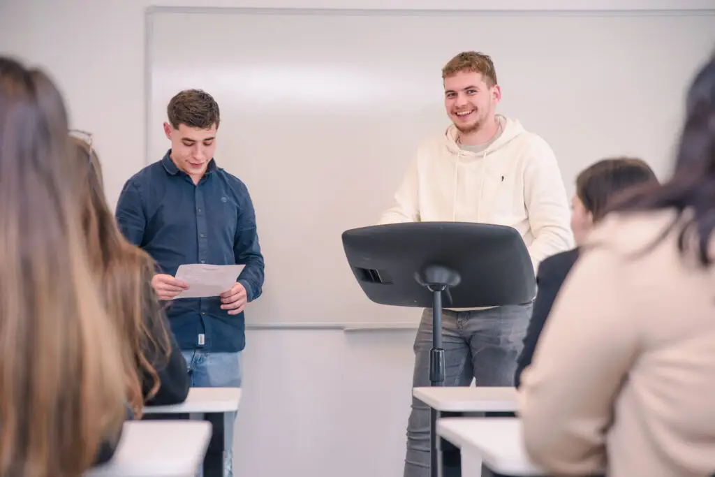 Photo de deux étudiants passant un oral devant le reste de leur classe.